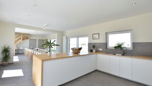 A modern kitchen with a wooden countertop, white cabinets, and a large island. There are potted plants, a window, and a staircase leading to another room.