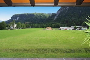A grassy field with a small building and houses in the background, surrounded by forested mountains.