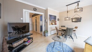 A modern living room with a flat-screen TV, a dining area with a round table and chairs, and a kitchen area with cabinets and a hanging light fixture.