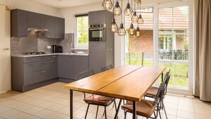A modern kitchen with grey cabinets, a wooden dining table, and a large window with a view of a brick house. - Holiday Villa with Garden (Bad Bentheim)
