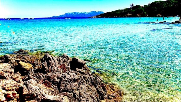 Eine felsige Küste mit klarem, blauem Wasser und Bergen in der Ferne.