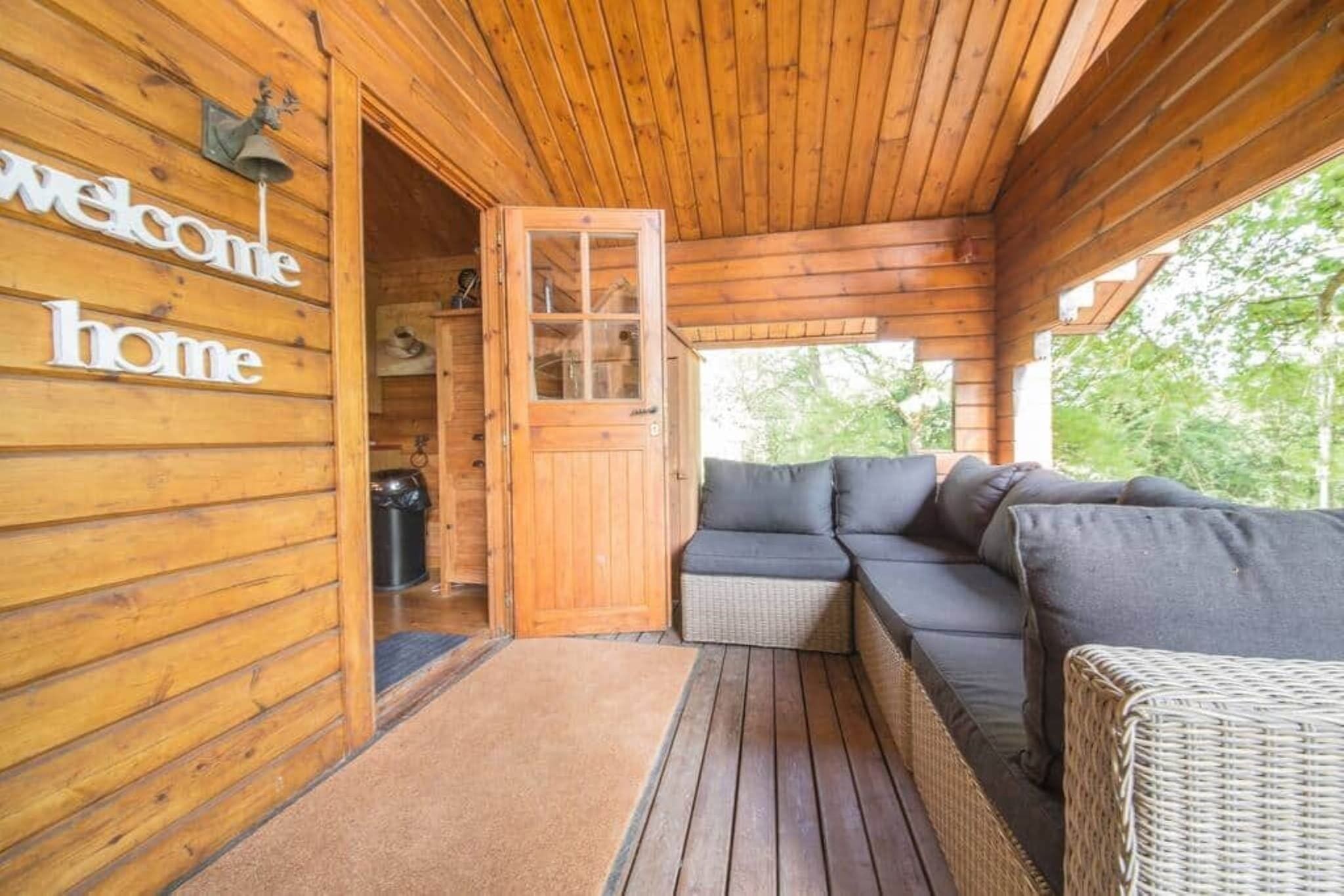 A wooden cabin with a 'Welcome home' sign, a glass door, a dark-colored sofa, and a wicker chair.