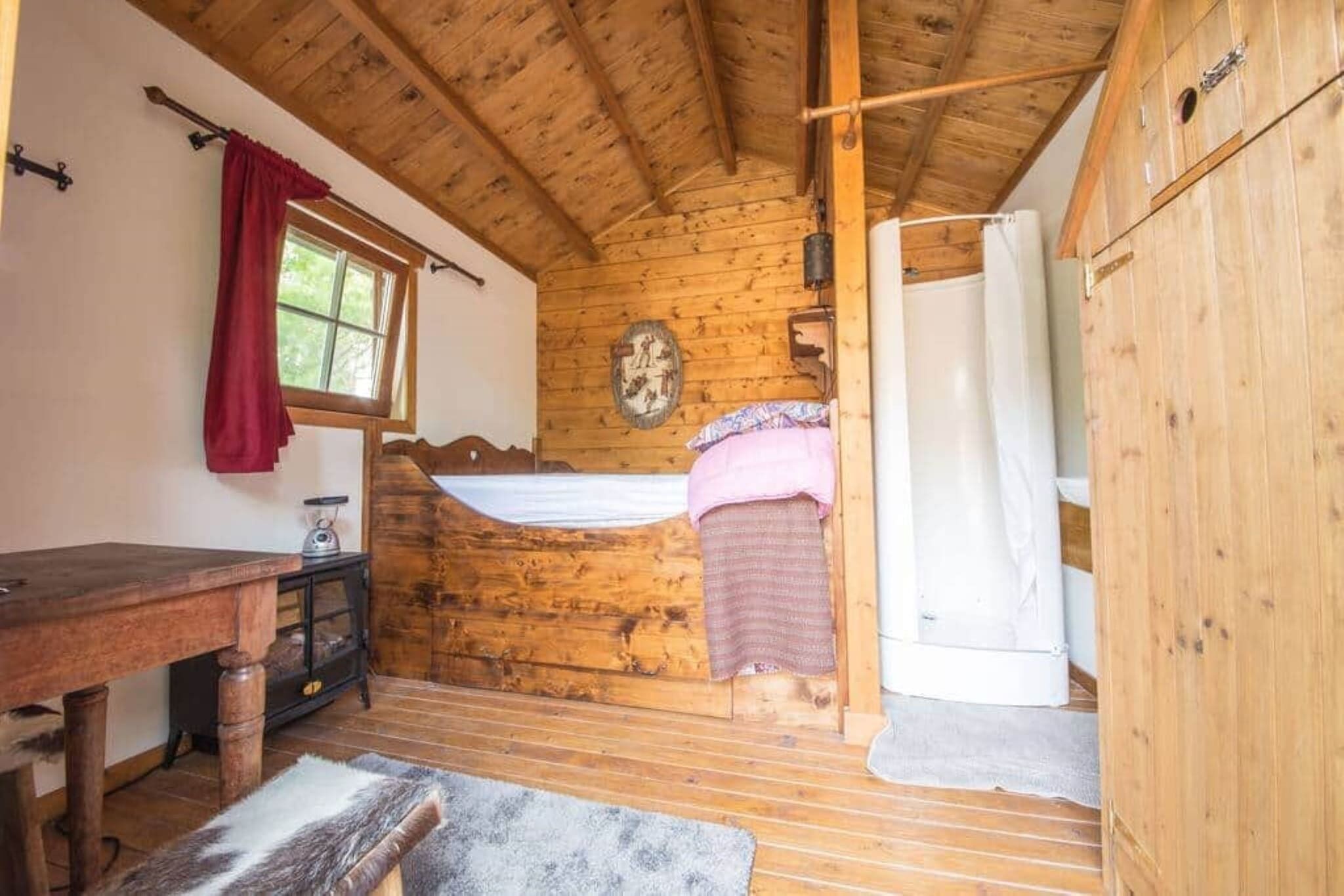 A wooden cabin room with a bed, a table, and a window with a red curtain.