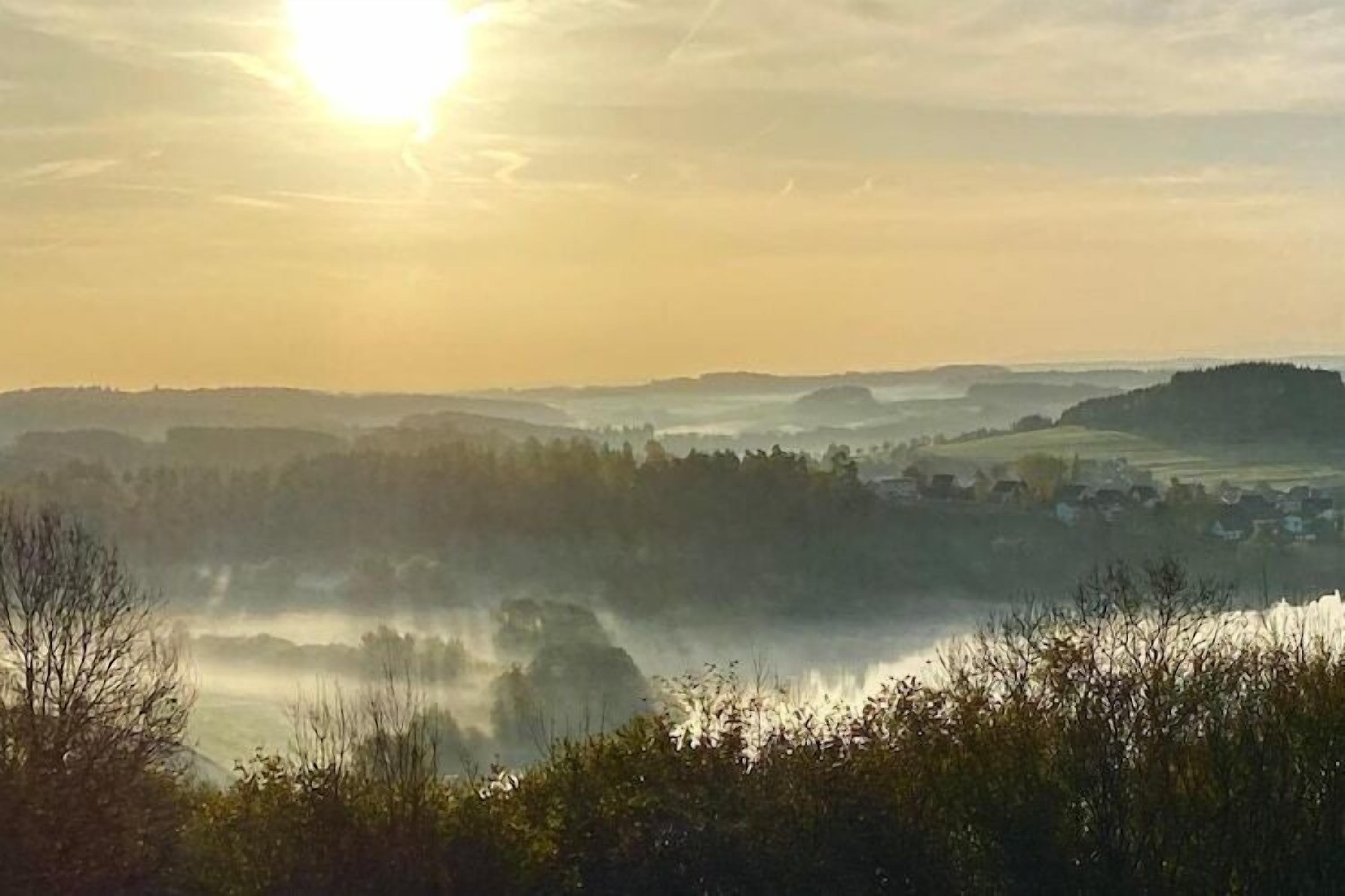 Un paysage brumeux avec une étendue d’eau, des collines ondoyantes et un petit village au loin.