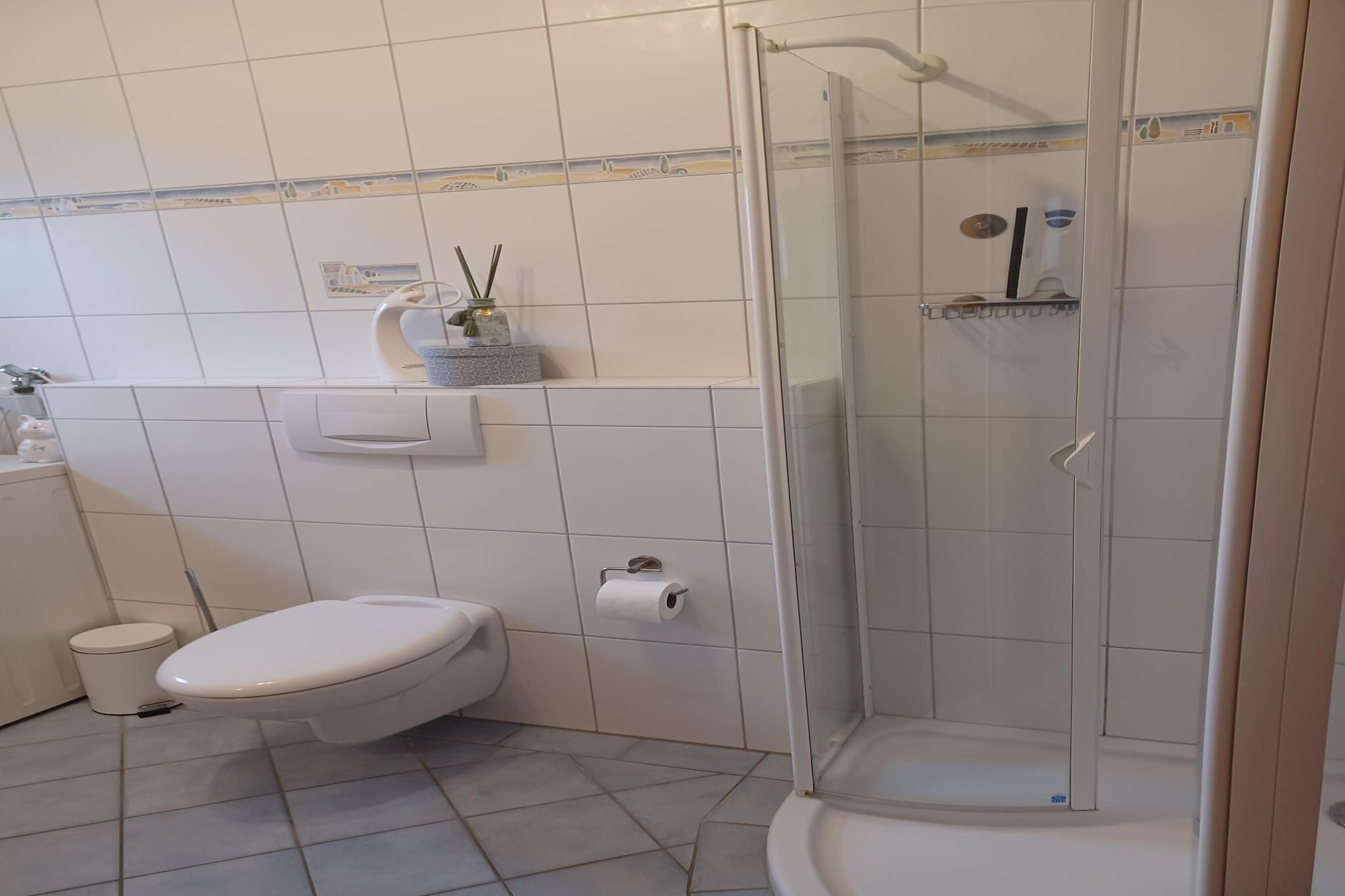 Ett badrum med en toalett, en liten papperskorg, en dusch med glasväggar och en hylla med toalettartiklar.