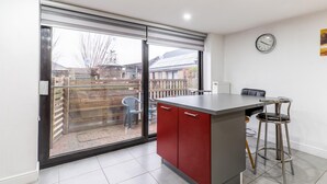 Une cuisine avec une porte-fenêtre coulissante donnant sur une terrasse, un meuble rouge, un tabouret de bar en acier inoxydable et une horloge au mur.