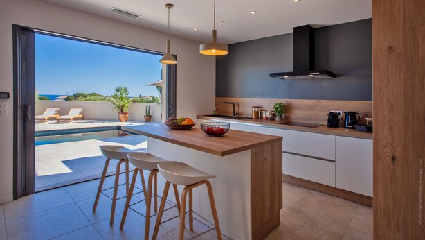 A modern kitchen with a large island, bar stools, and a view of a pool and outdoor seating area. - Superb villa with pool and sea view (Monticello)