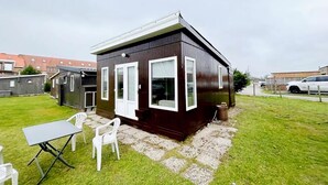 A small, dark wooden house with white-framed windows and a door, surrounded by a grassy yard with a table and chairs. - Lovely holiday home in Vlaanderen with garden (Vlaanderen)