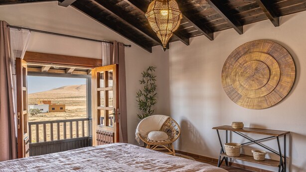 Ein Schlafzimmer mit einem großen Bett, einem Korbstuhl, einem Nachttisch aus Holz und Blick auf eine Wüstenlandschaft.