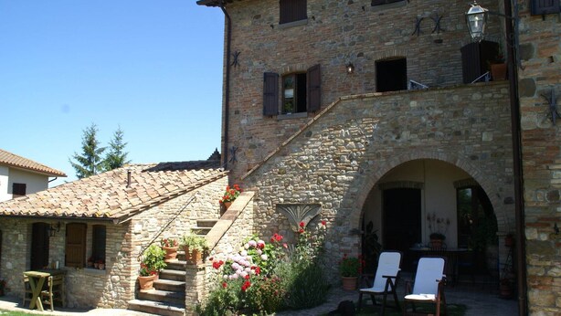 A stone building with a staircase leading to an upper level, a covered outdoor seating area with chairs, and a small garden with potted plants.