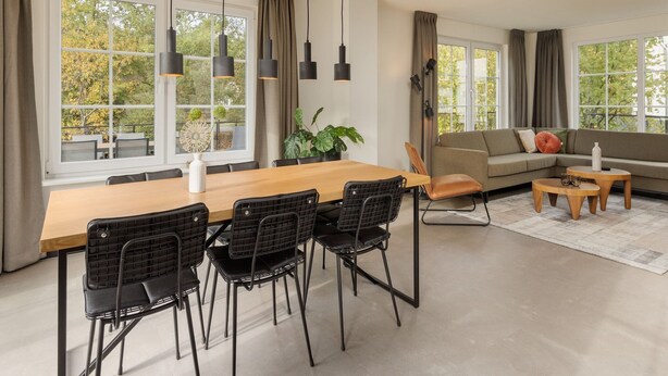 A modern living room with a wooden dining table, black chairs, a sofa, and a coffee table. Large windows with curtains provide natural light and views of trees. - Holiday Home in Bad Bentheim with Sauna (Bad Bentheim)