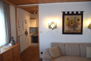 Uma sala de estar acolhedora com teto de madeira, um sofá bege, um quadro de girassóis na parede e uma área de cozinha ao fundo.