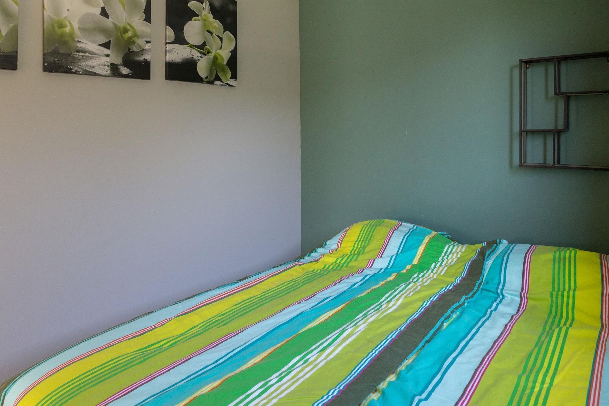 Una cama con un edredón a rayas de colores, una pared con tres cuadros de flores blancas y una ventana con marco de metal.