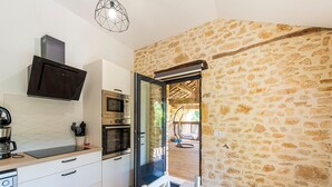 Une cuisine moderne équipée d’un four à micro-ondes, d’un four et d’une machine à café encastrés. On y trouve une table à manger avec des chaises et une bouteille de vin. La cuisine donne sur un mur en pierre et sur un balcon où est suspendue une chaise.