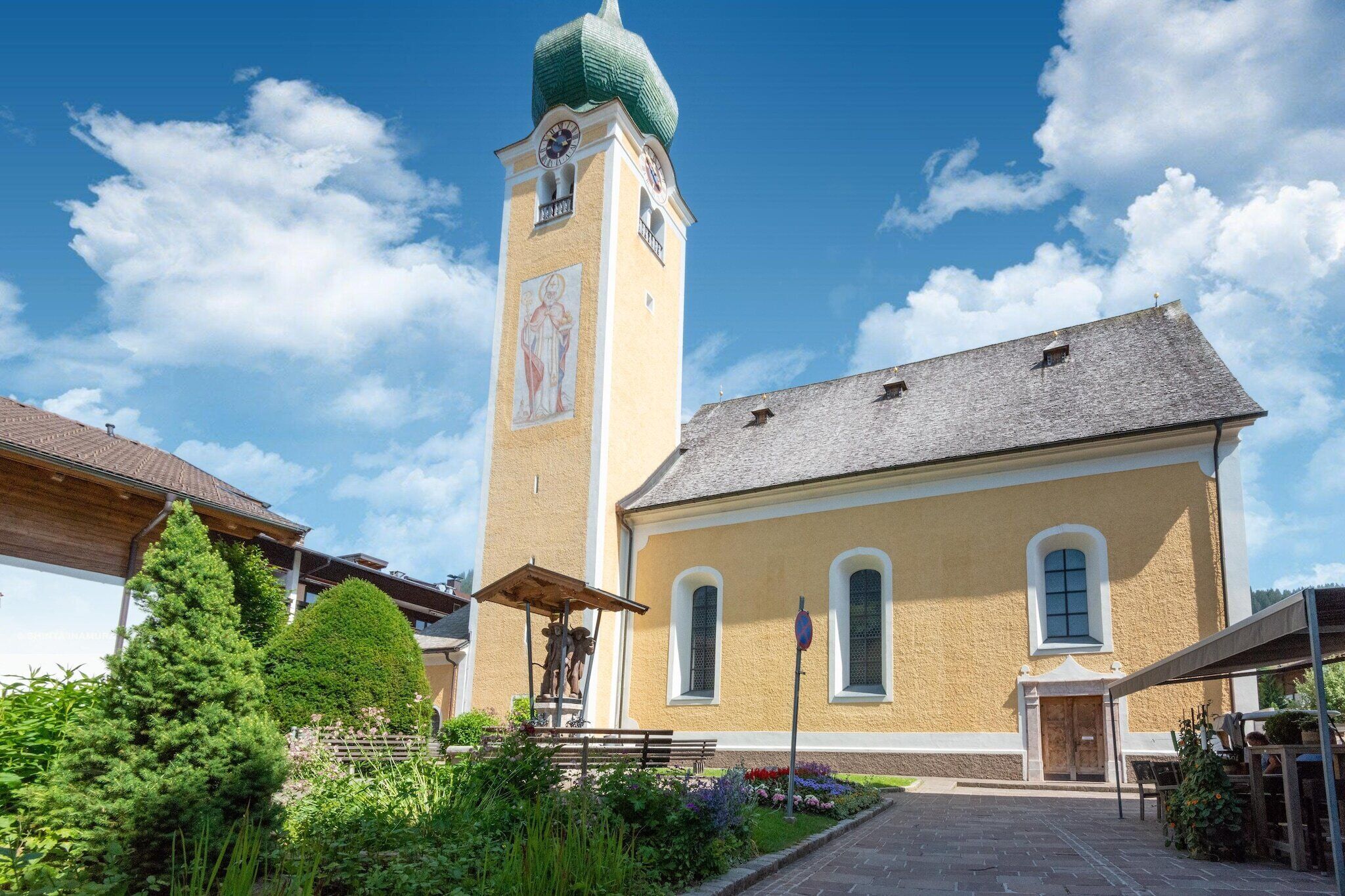 A church with a tall tower and a clock, surrounded by greenery and a paved walkway.