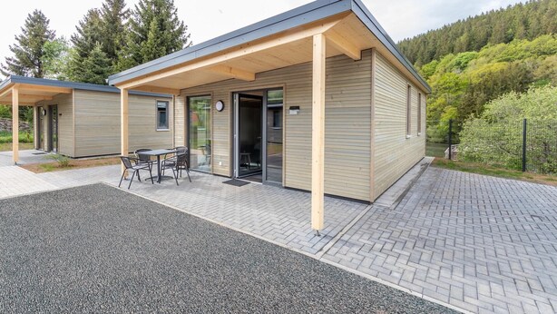 A modern building with a covered patio area, a small outdoor seating arrangement, and a view of a forested hillside. - Komfortables Bungalow am Kronenburger See mit Terrasse (Hallschlag)