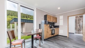 A modern kitchen with wooden cabinets, a microwave, and a built-in oven. There is a dining area with a table and chairs, and a large window offering a view of a forested area. - Komfortables Bungalow am Kronenburger See mit Terrasse (Hallschlag)