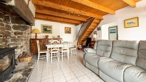 A cozy living room with a stone fireplace, wooden ceiling beams, a dining area with white chairs, and a grey sofa. - Stone-house cottage in Saussey with garden (Saussey)