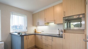 A compact kitchen with light wood cabinets, a built-in oven, and a window with blinds. - Ground floor apartment with large garden (Middelkerke)