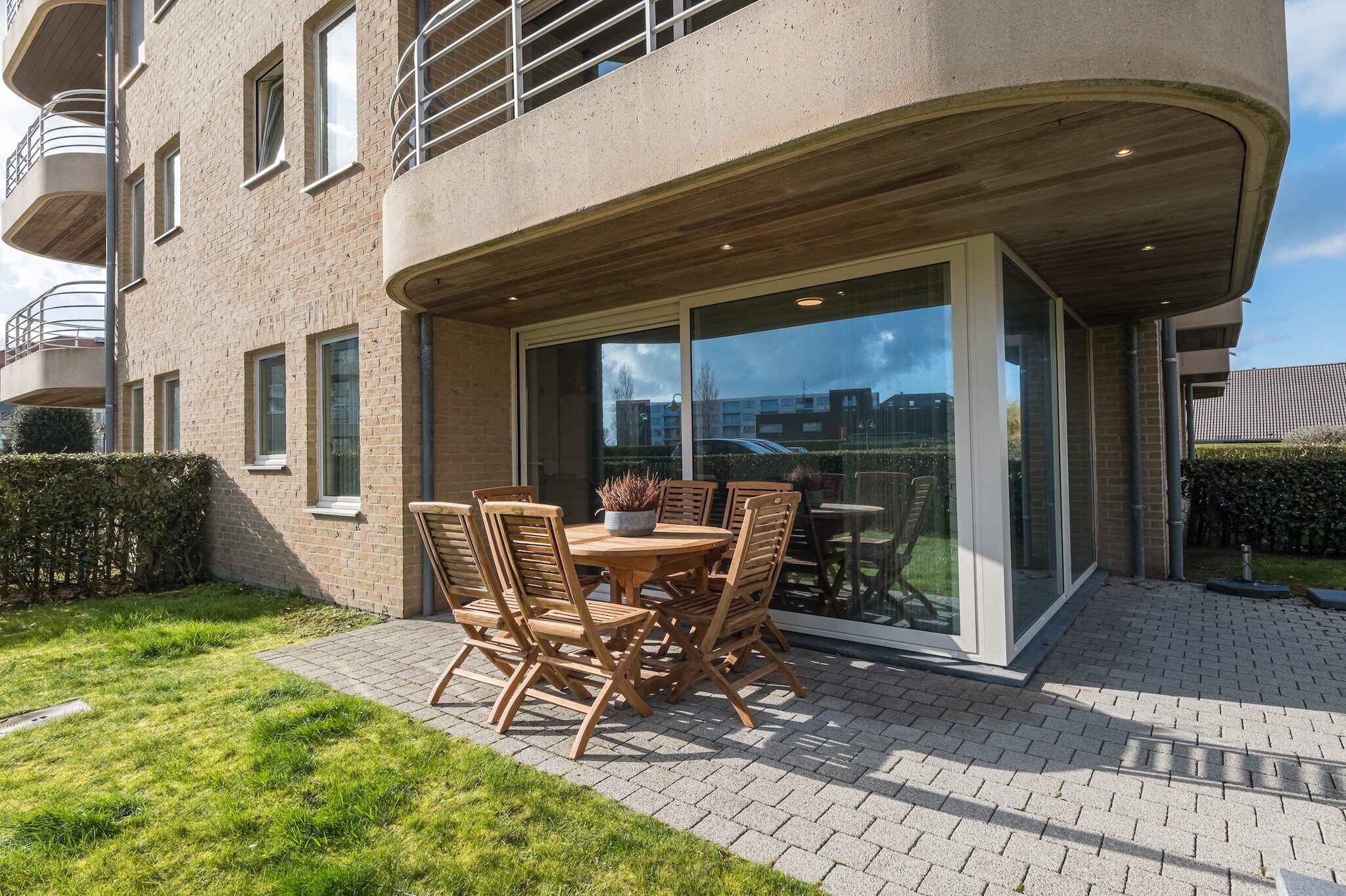En moderne lejlighed med en patio, der har et rundt træbord og stole, en potteplante og en glasdør, der fører ud til en balkon.
