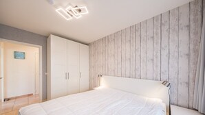 A bedroom with a white bed, a wooden wall panel, and a ceiling light fixture. - Ground floor apartment with large garden (Middelkerke)