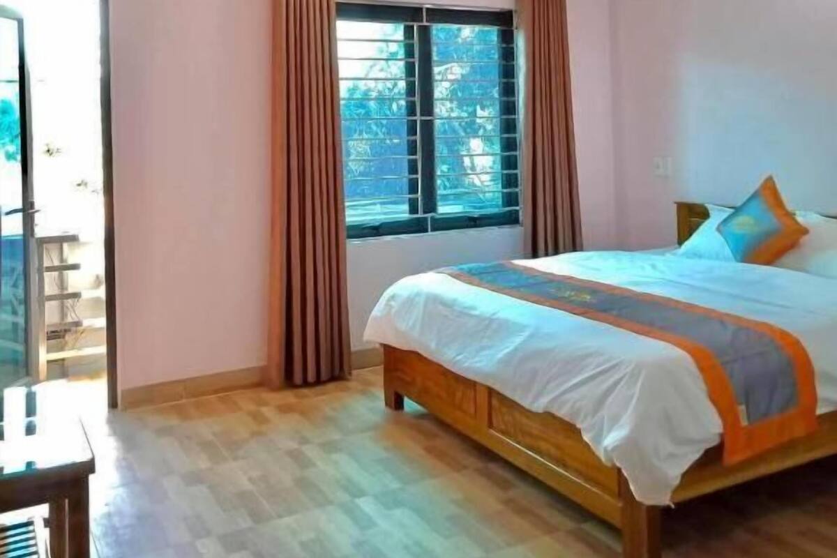 A bedroom with a large bed, a window with curtains, a ceiling light, and a wooden floor.