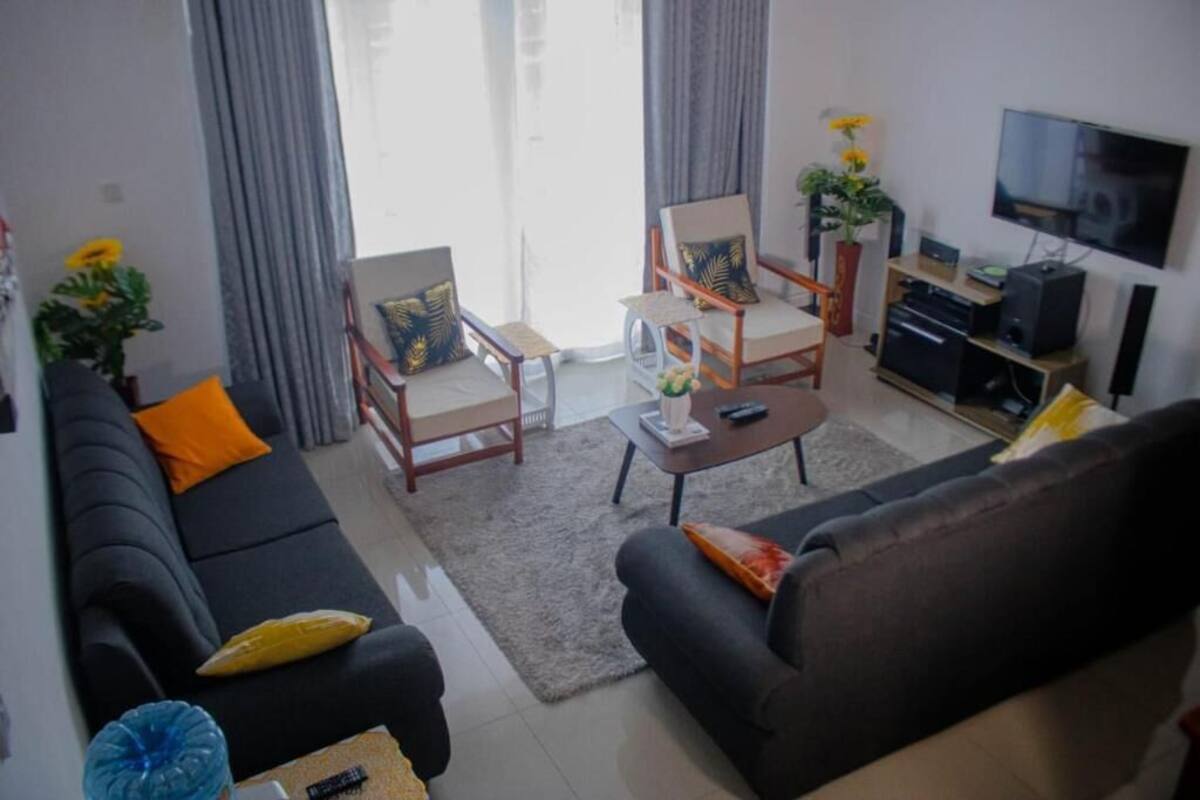 A modern living room with a black sofa, a coffee table, and two armchairs. A flat-screen TV is mounted on the wall, and there are potted plants on the windowsill.