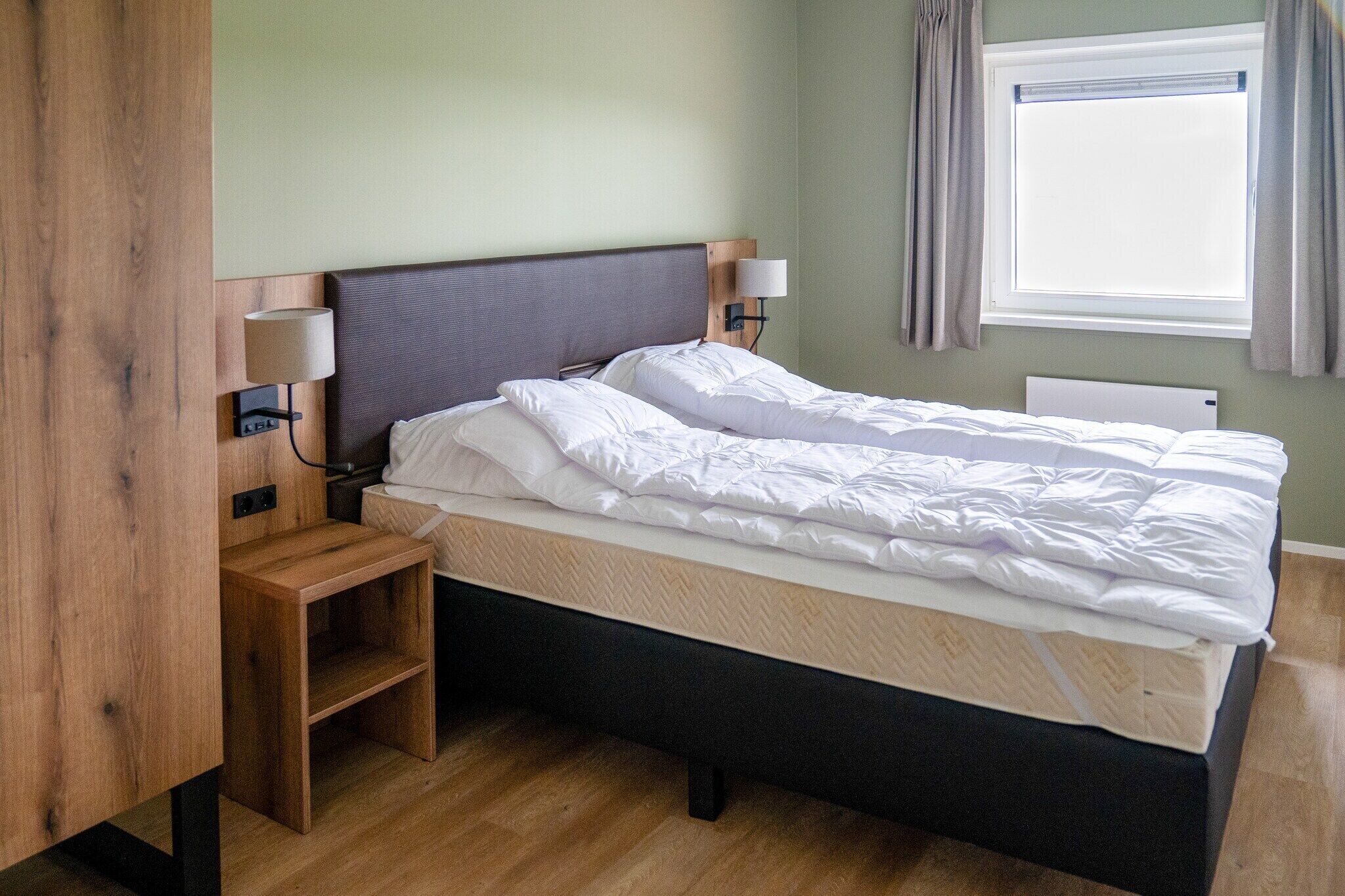 Una cama bien tendida con cabecera, lámparas de noche, mesita de noche de madera y una ventana con cortinas.