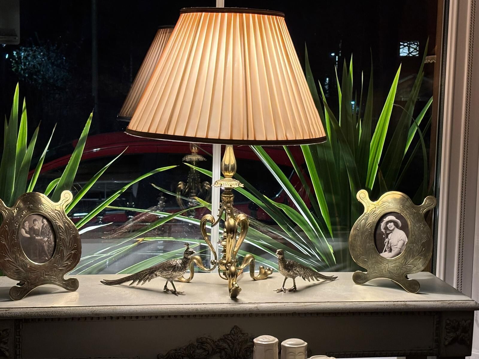 A lamp with a pleated shade, two ornate bird figurines, and two framed pictures on a mantelpiece.