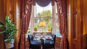 A corner room with a dining table set for two, a potted plant, and a window with curtains. - Brooklands Guest House  (Penrith)