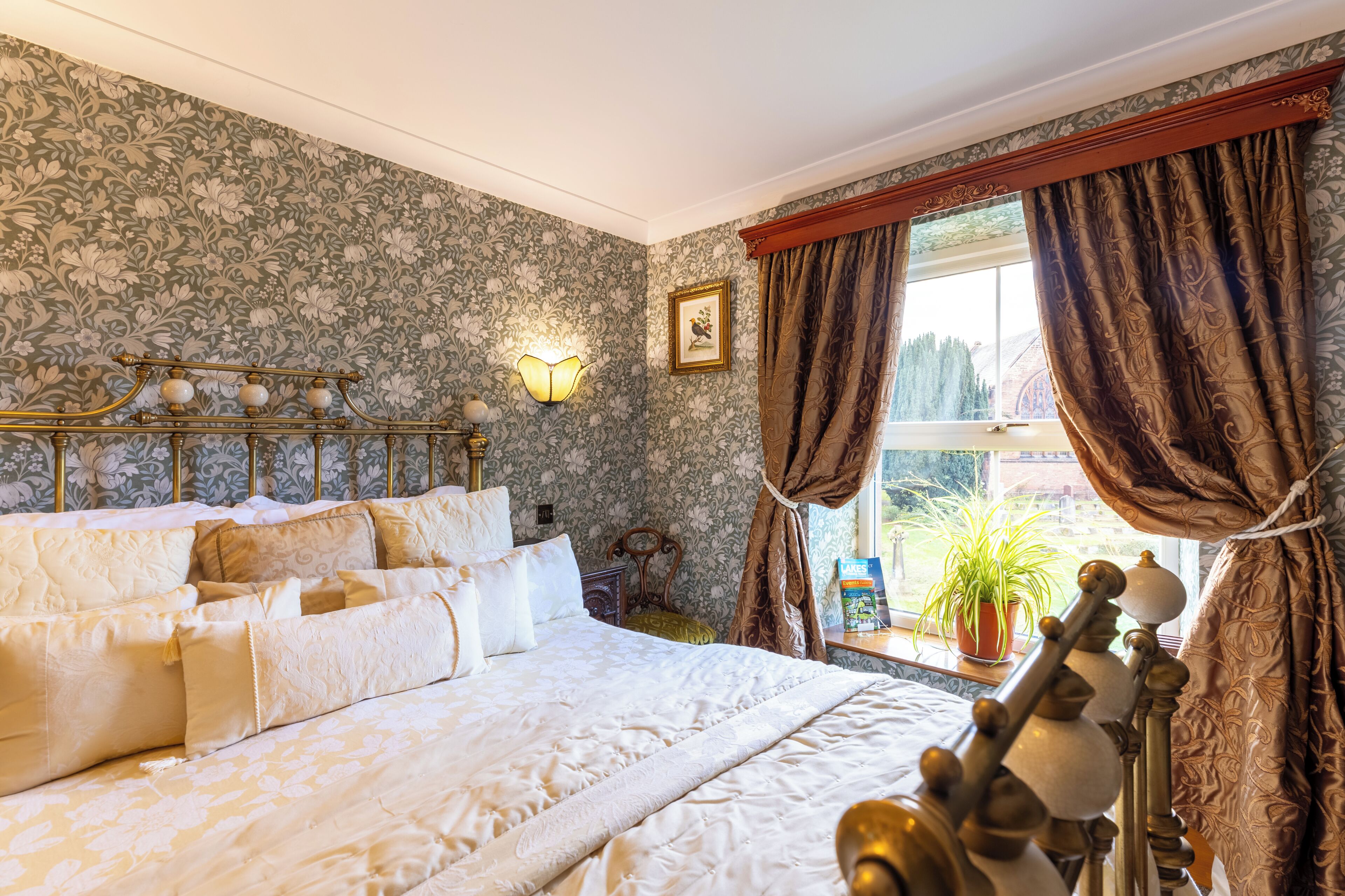 Une chambre avec un grand lit, un papier peint à motifs floraux, un luminaire fixé au mur, une fenêtre avec des rideaux et une plante en pot près de la fenêtre.