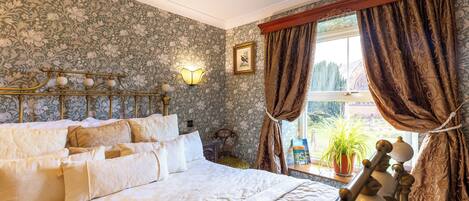 Une chambre avec un grand lit, un papier peint à motifs floraux, un luminaire fixé au mur, une fenêtre avec des rideaux et une plante en pot près de la fenêtre.