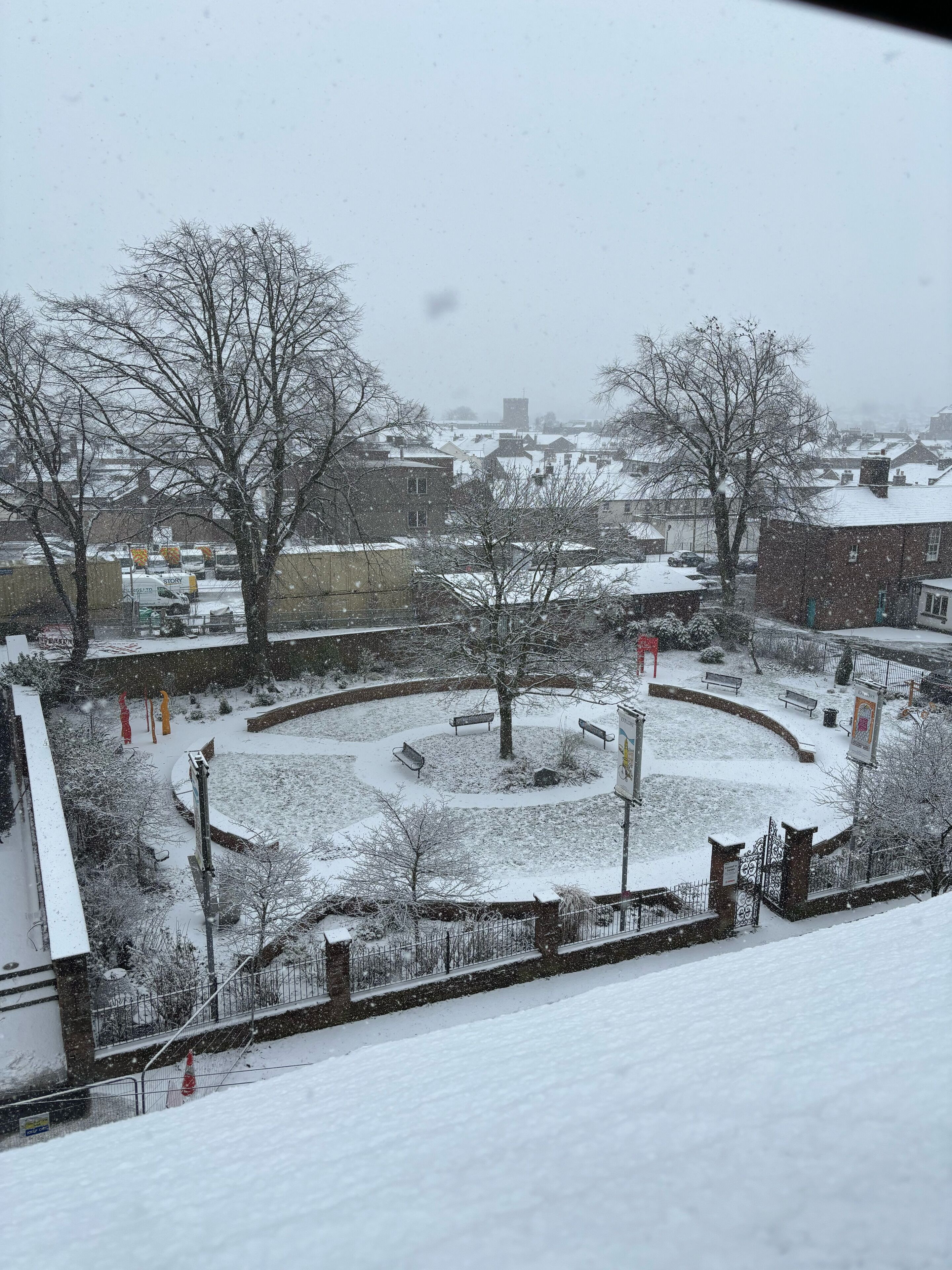 A snow-covered courtyard with benches and a central tree, surrounded by buildings and a fence.