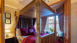 A four-poster bed with a red bedspread and multiple pillows, a nightstand with a lamp, and a window with curtains. - Brooklands Guest House  (Penrith)