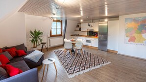A modern living room with a sofa, a dining table with chairs, a small side table, a potted plant, and a kitchen area with cabinets and appliances. - Elegante Ferienwohnung mit Außenpool Schwarzwald (Herrischried)