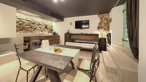 A modern kitchen with a stone wall backsplash, a dining area with a table and chairs, a built-in bench, and a flat-screen TV mounted on the wall. - BASTINICA KRK Studio Ap 1, OldTown, CityCenter (Krk)
