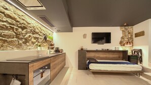 A modern bedroom with a stone wall, a bed, a TV, and a built-in kitchen area. - BASTINICA KRK Studio Ap 1, OldTown, CityCenter (Krk)