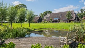 Een traditioneel huis met rieten dak, een vijver en een houten terras met een bank.