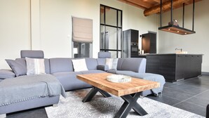 Een moderne woonkamer met een grijze modulaire bank, een houten salontafel en een keukenhoek met donkere kasten en een inbouwoven.