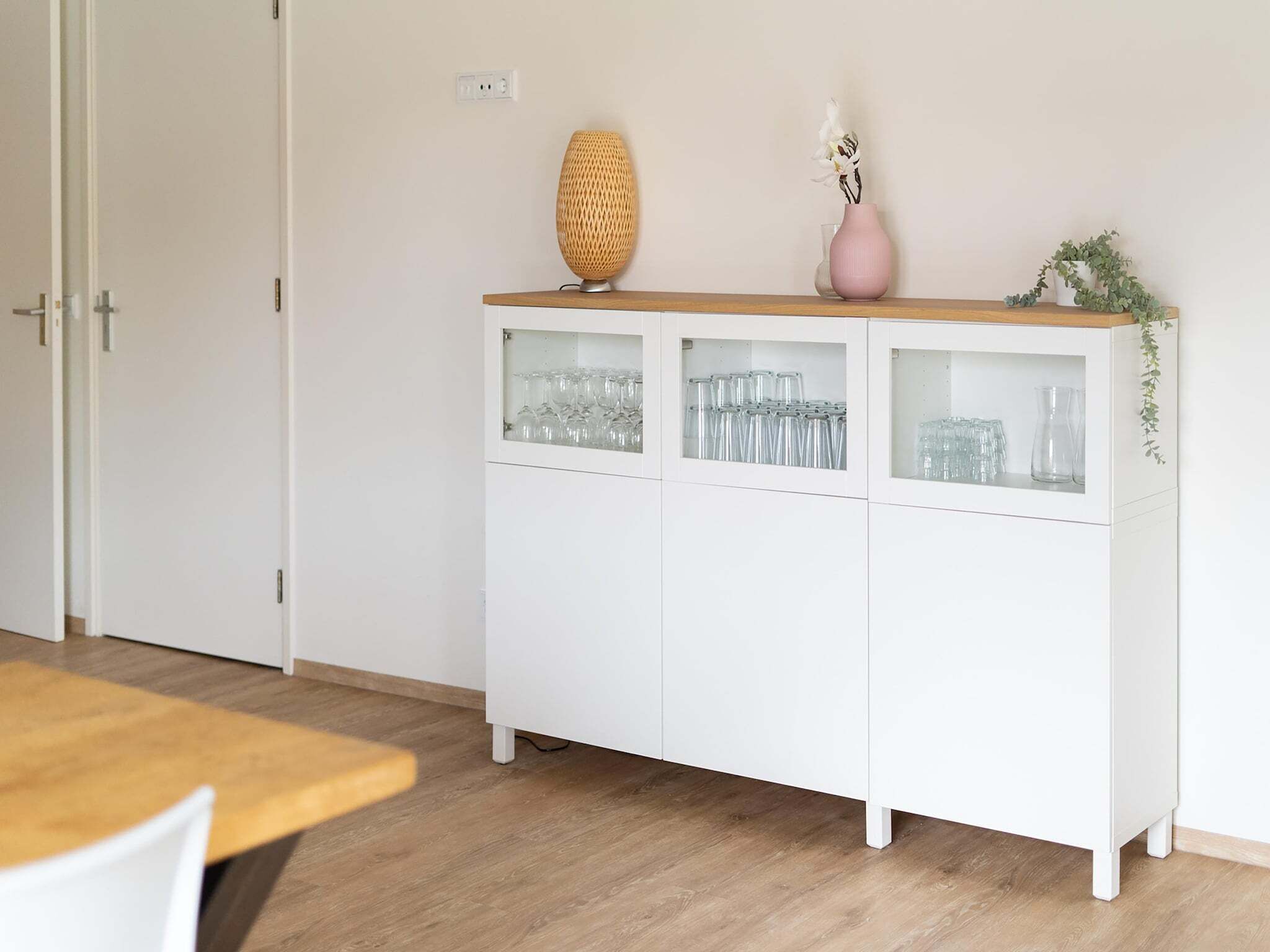 A white cabinet with glass-fronted shelves containing glassware, a woven lamp, and a pink vase with white flowers.