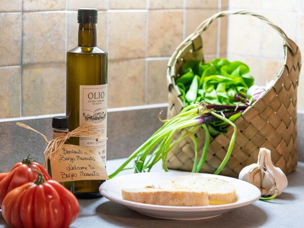 A kitchen counter with a bottle of olive oil, a basket of greens, a tomato, a garlic bulb, and a slice of bread.