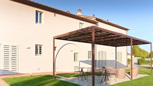 Un área de estar al aire libre con techo, mesa y sillas, una estructura de pérgola y un edificio blanco con contraventanas y ventanas.