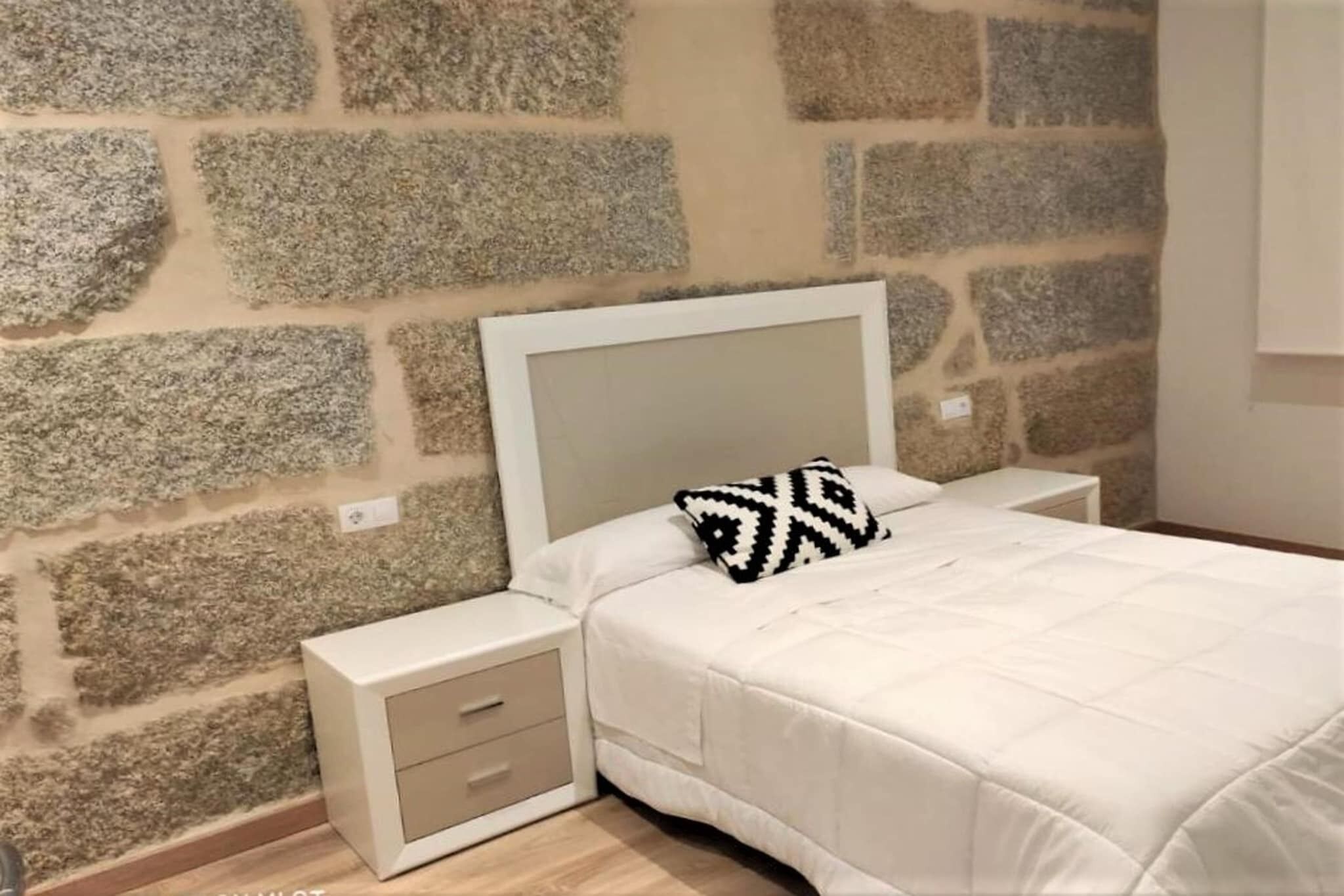 Un dormitori amb una paret de pedra, un llit amb capçalera blanca, una tauleta de nit amb calaixos i un coixí estampat.