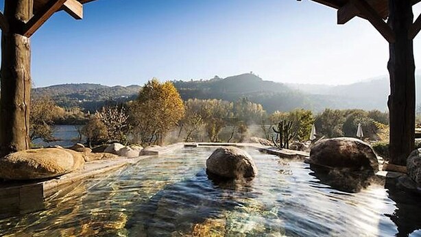 Piscina cristalina con rocas, rodeada por una estructura de madera y vistas a colinas.