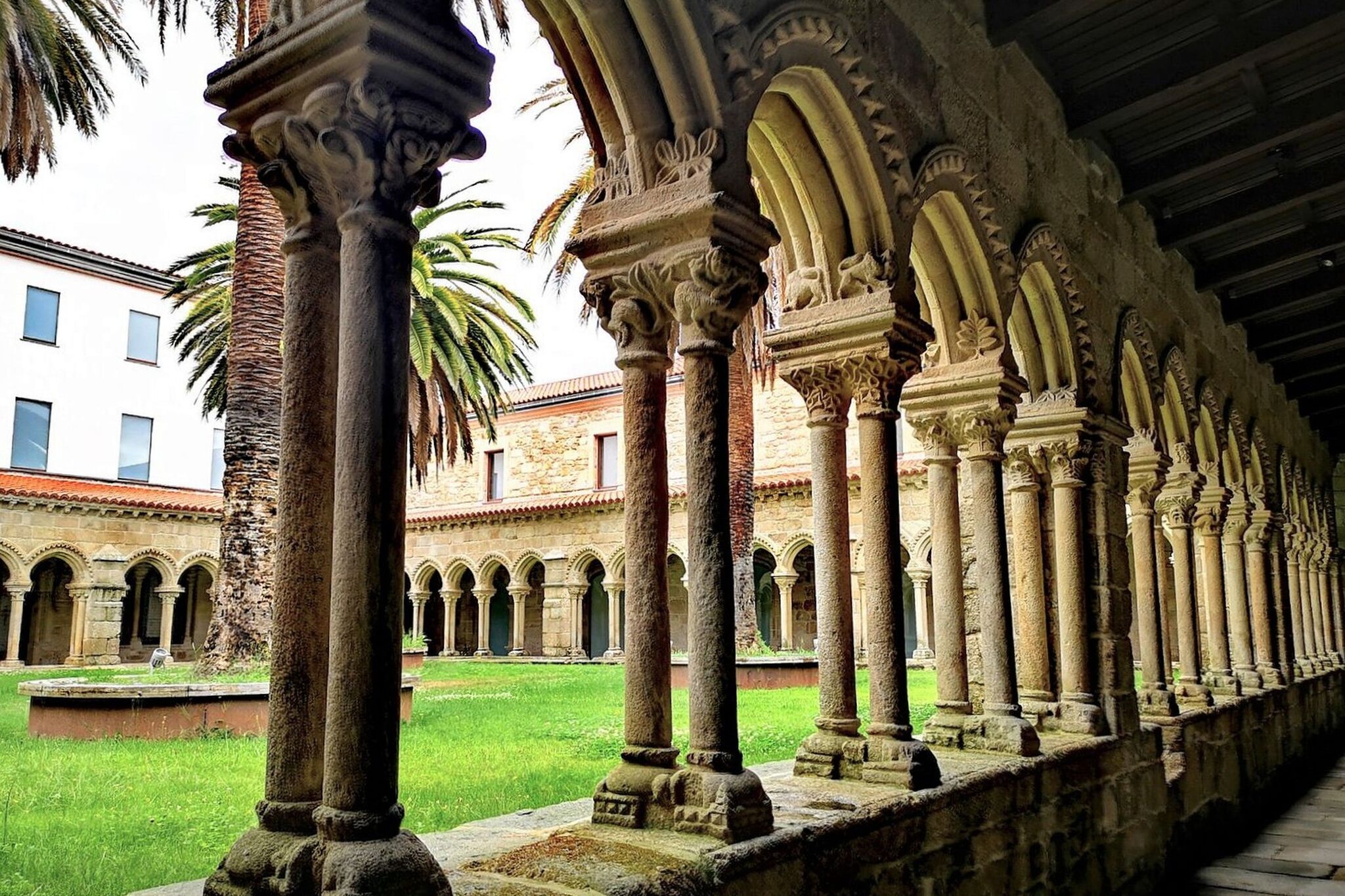 A courtyard with a series of columns and arches, a grassy area, and a building in the background.