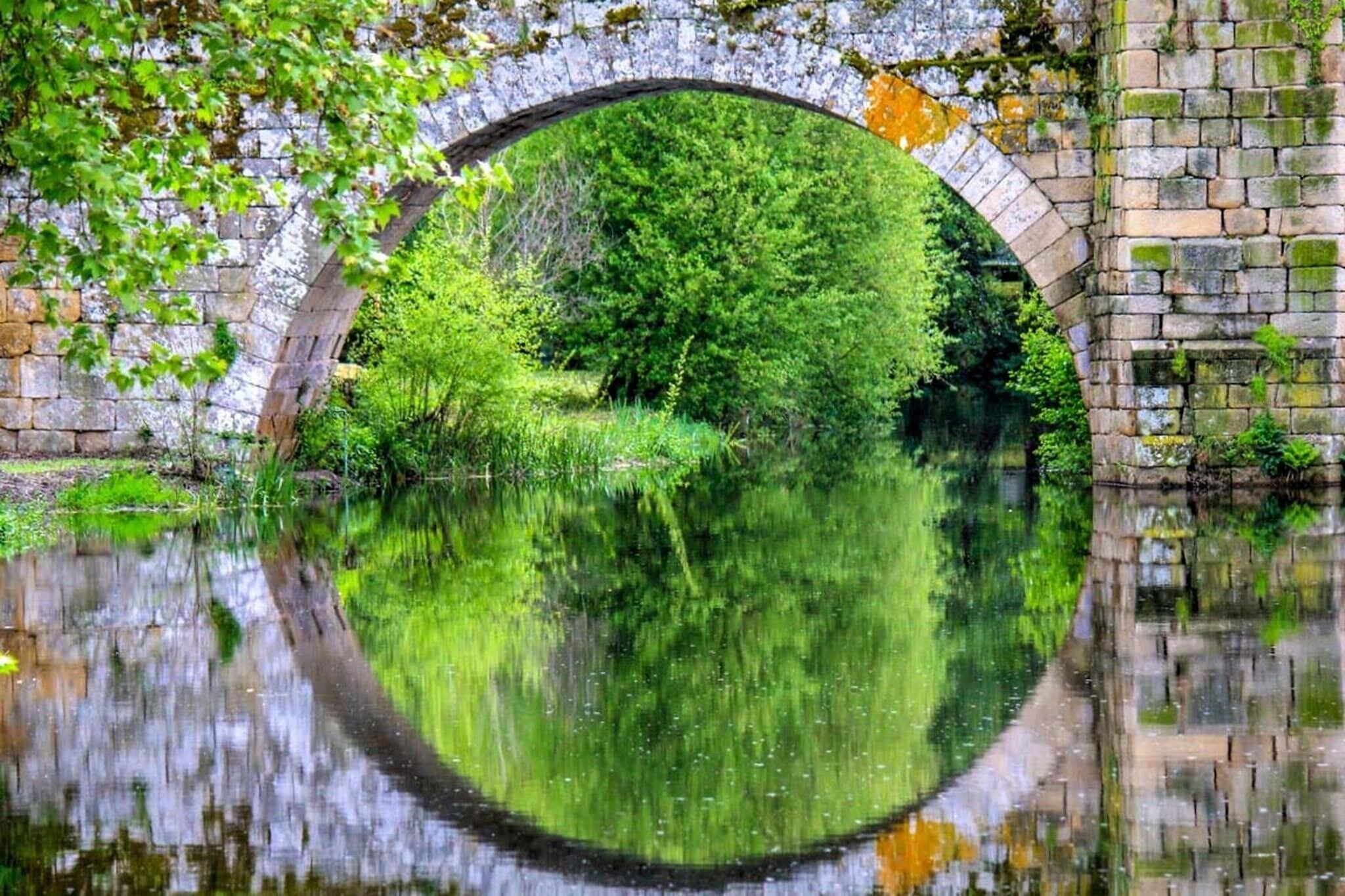 A stone bridge arching over a calm river, surrounded by lush greenery and trees.
