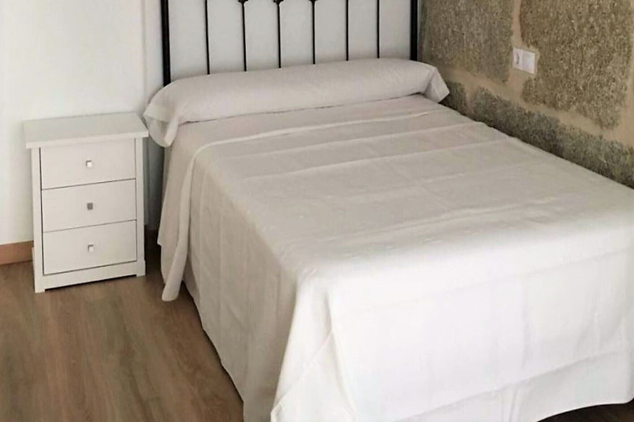 Quarto individual com uma cama branca, mesa de cabeceira com gavetas e parede de pedra.