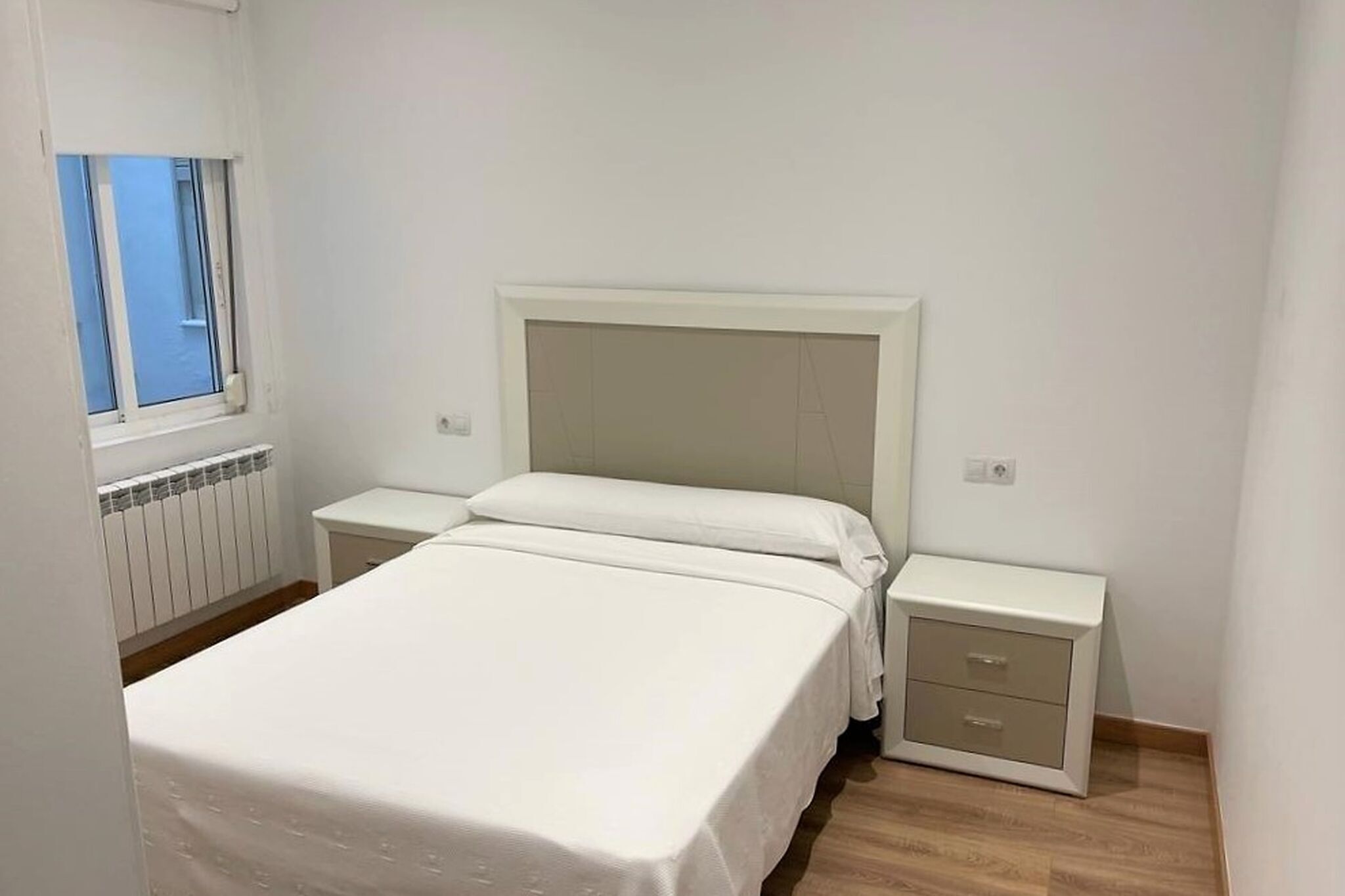 Une chambre à coucher avec un lit, deux tables de chevet, une fenêtre et un radiateur.