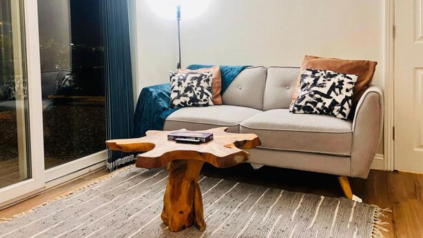 A modern living room with a grey sofa, two patterned cushions, a wooden coffee table, and a large window with a view of the outdoors. - Lolite Apartment in Cambridge (Cambridge)