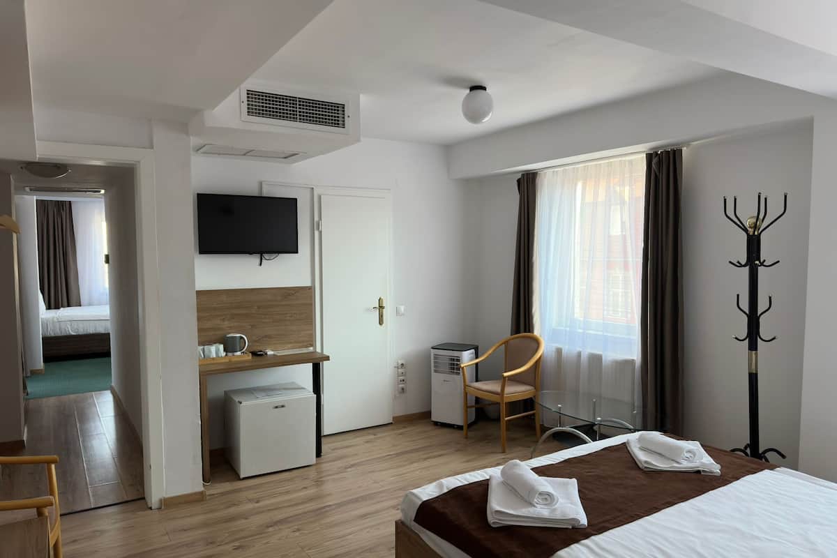 Ett hotellrum med en säng, ett skrivbord, en stol, en tv, en klädställning och ett fönster med gardiner.